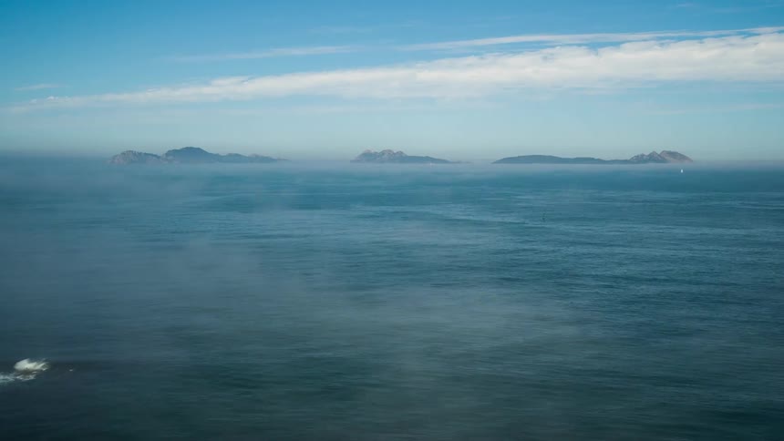 Fog moves across water and rock formations are seen in the distance