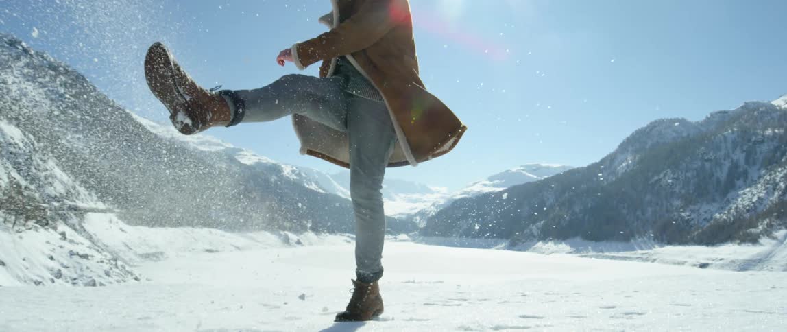 Person standing in snowy field backed by mountains kicks at snow