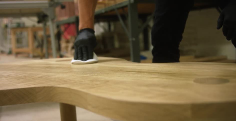 Man wearing black gloves applies varnish over a wooden coffee table inside a workshop