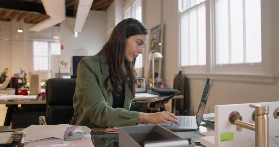 A woman looks for a piece of paper and types information from it onto her laptop