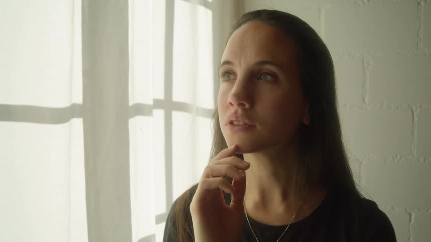 Young woman thinking while standing near white curtains on a white room
