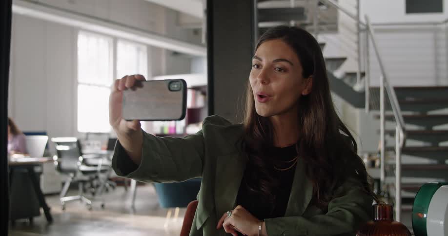 A woman waves while sitting at her desk at work on a video call on her smartphone