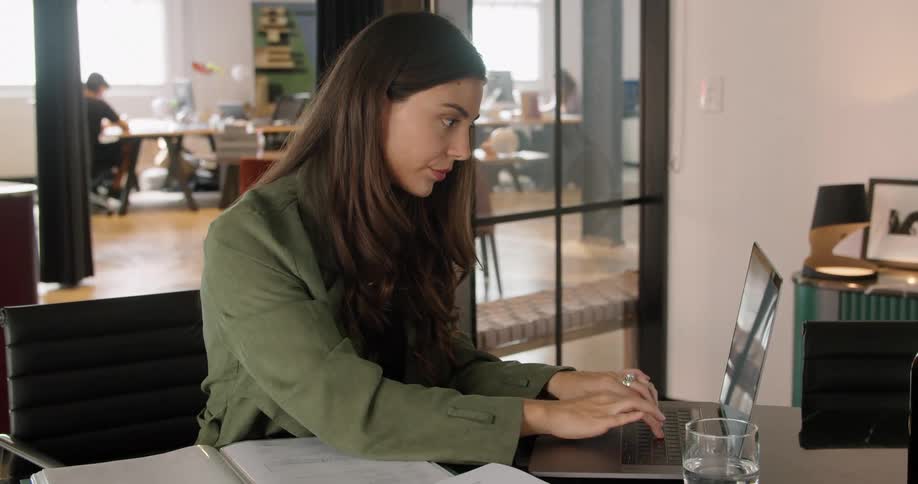 A woman sitting at a desk in an office looking over paperwork scattered on the desk and typing on a silver laptop computer at daytime