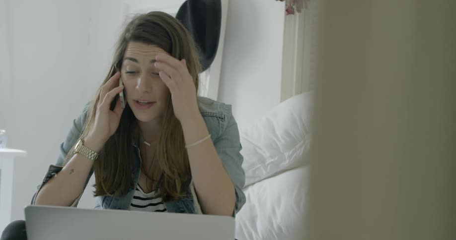 A woman sits in front of a laptop in a bedroom talking on the phone