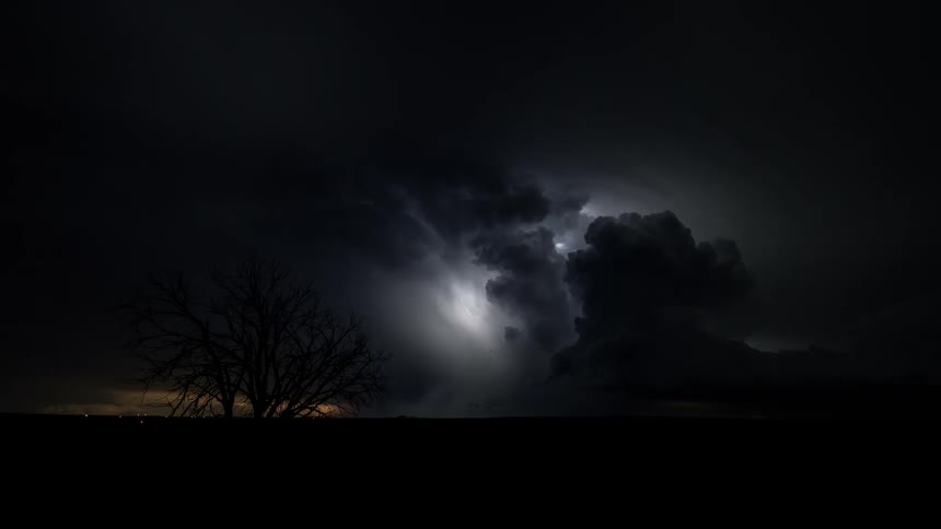 Lighting flashes brightly from storm clouds in the night sky near a bare tree