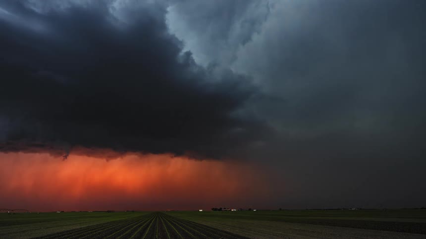 Traffic flows toward lit building on edge of crop fields as storm front roils overhead