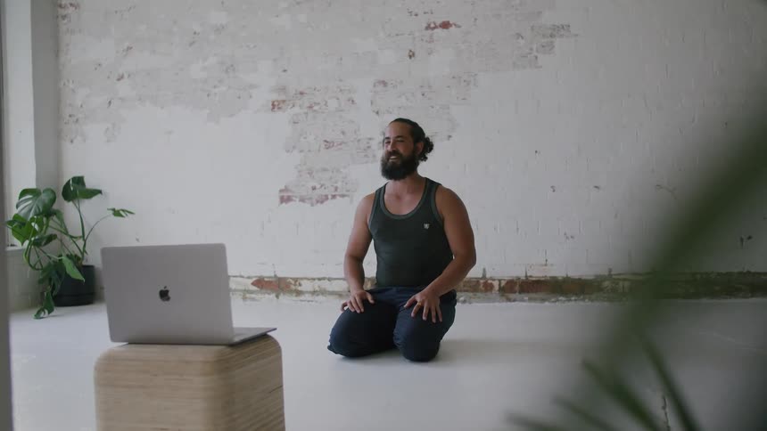 A man kneeling on the floor waving to a laptop with a wall and a plant in the background