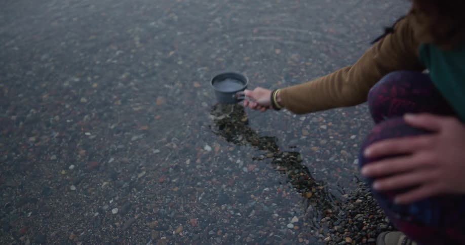 Woman scooping up water with metal cup at shoreline on a lake