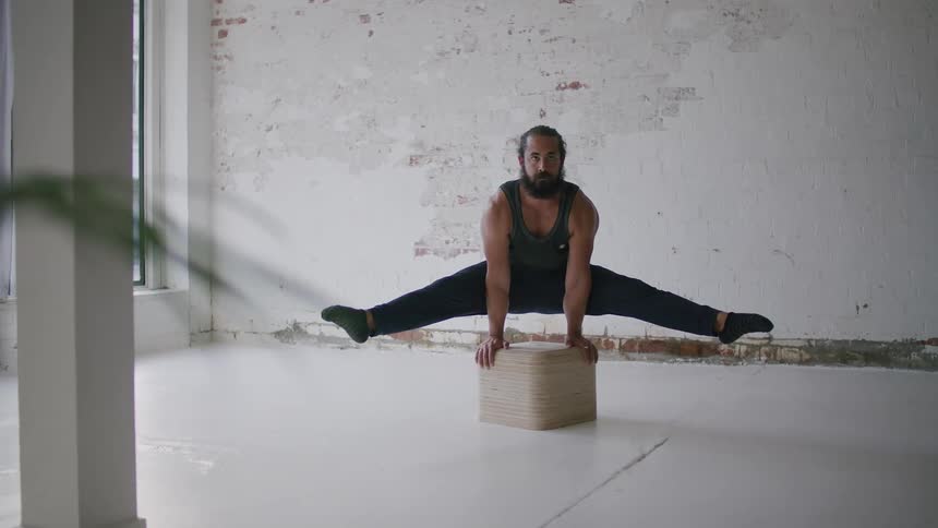 Bearded man wearing sports clothes doing yoga balances over a box inside a room