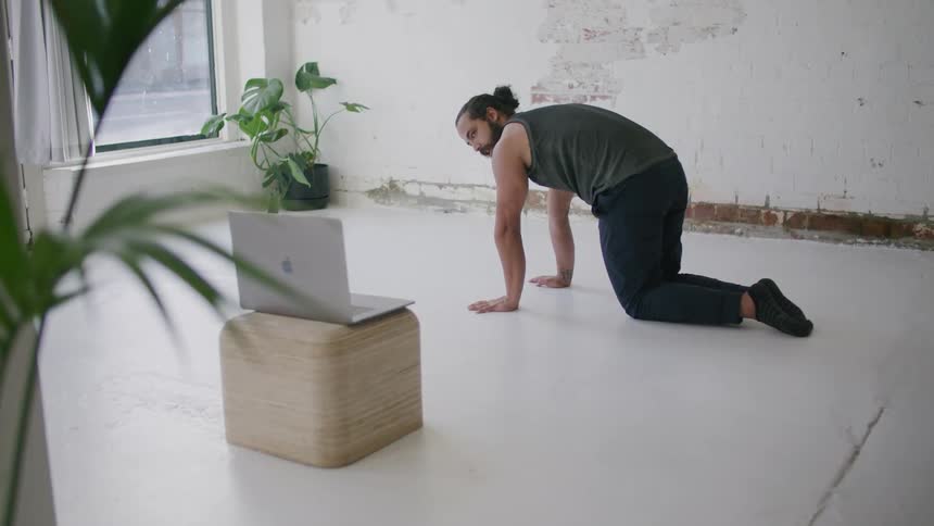 A man kneeling on the floor talking to a laptop with a wall and a plant in the background