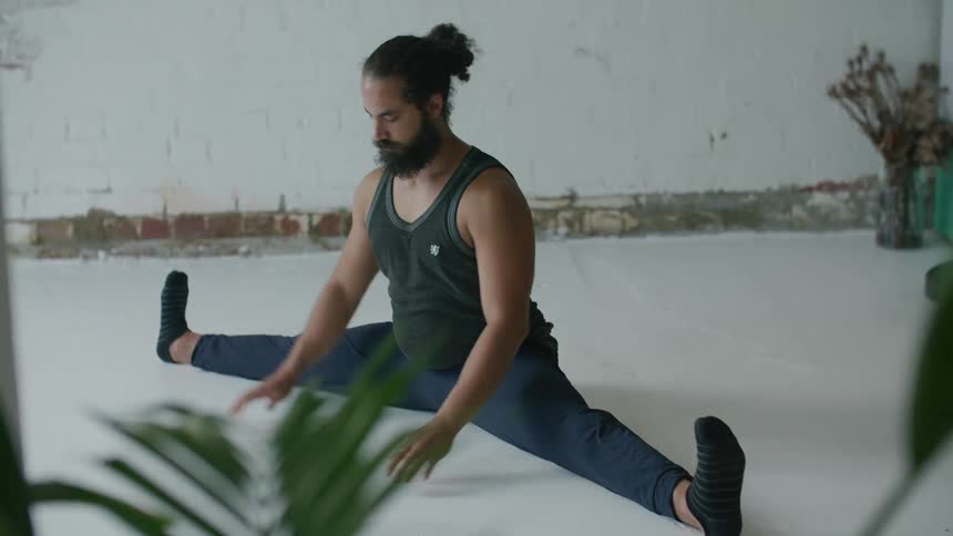 A man stretches while sitting on the ground in splits while surrounded by plants