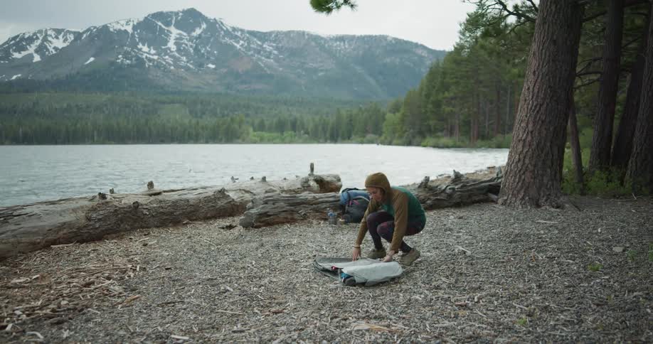 A young woman unpacks a tent on a campground next to a lake in the wilderness on a cloudy day