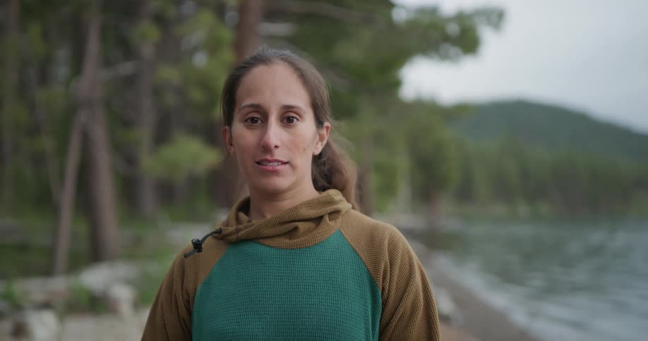 A woman stands outdoors in a forest next to a lake smiling for a picture towards the camera