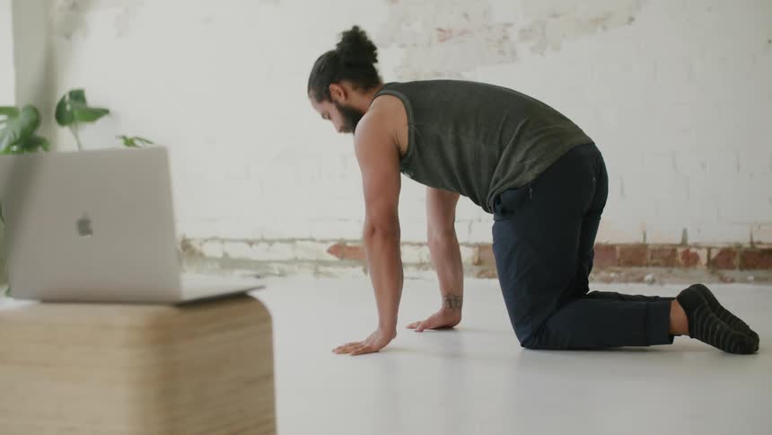 A man on the floor stretching with a laptop in the foreground and a plant and a wall in the background