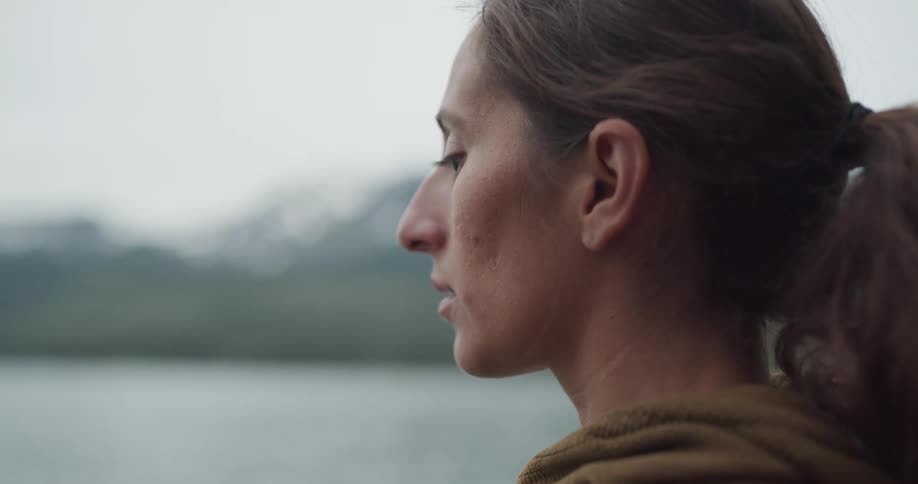 Side view of woman with a ponytail and a brown sweater standing and watching a lake on a cloudy day