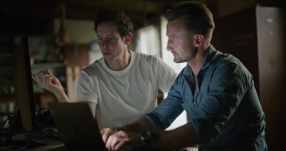 One man types on a laptop as another man points at a computer screen and they talk