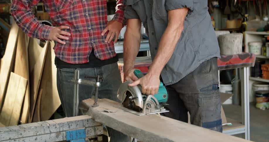 Father teaching son how to use power tools to cut lumber in the workshop