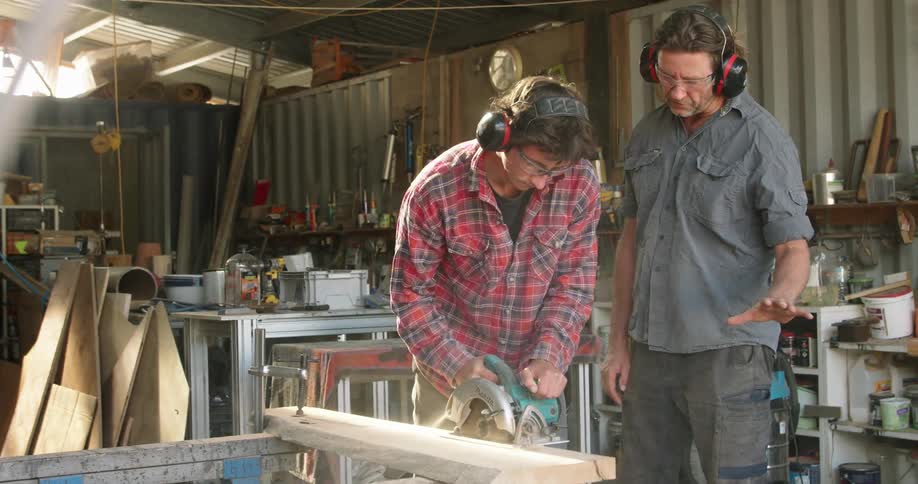 Two men in a workshop with goggles and earmuffs using an electric saw to cut wood