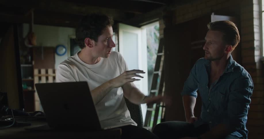 A man sitting before a laptop at a desk speaks with another man sitting next to him indoors during the day