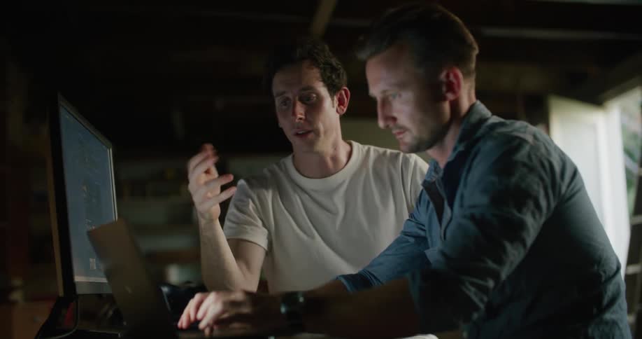 Two men sitting indoors at a table with a desktop screen and a laptop while one types and the other points