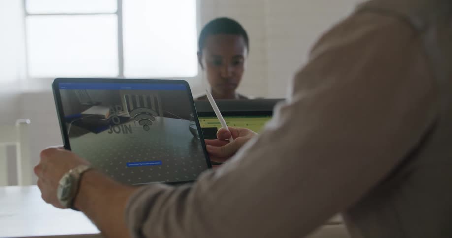 A man and a woman sitting at a table with laptops with a wall and a window in the background