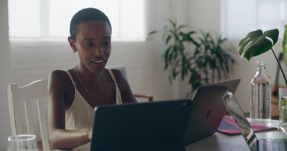 A woman sitting on a chair at a table in front of laptops with a wall and window in the background