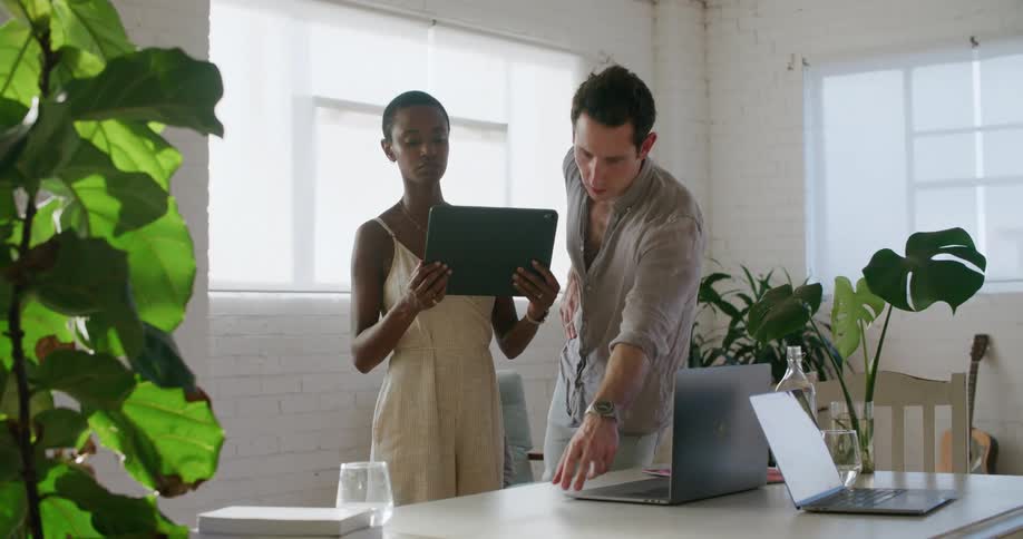Man with a shirt and jeans stands next to woman with short hair and a white dress holding a tablet in a room with white walls