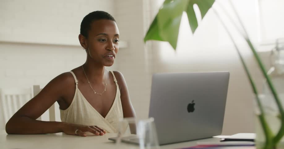 A woman sitting on a chair at a table talking to a laptop with a wall and window in the background