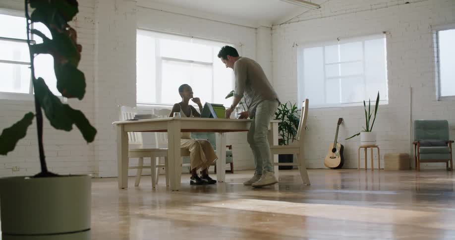 A man walking up to a table and sitting down where a woman sits with a laptop with a wall and window in the background