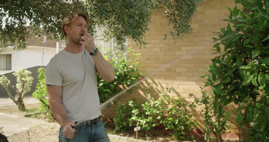 Muscular fit man watering plants ion his backyard garden at daytime