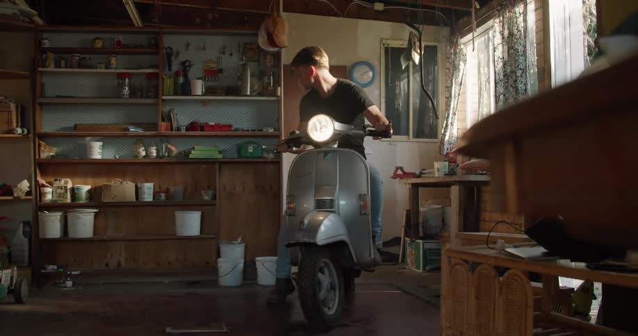 A man in a garage riding a moped with shelves and buckets in the background