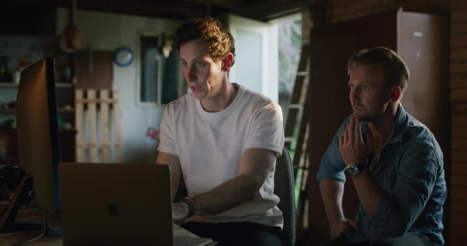 Two men in a room sitting at a table in front of a monitor and a laptop with a wall and door in the background