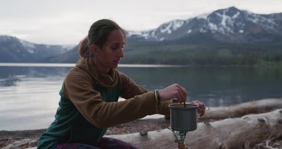 A woman by a lake sitting on a log with a mountain range the sky and clouds in the background