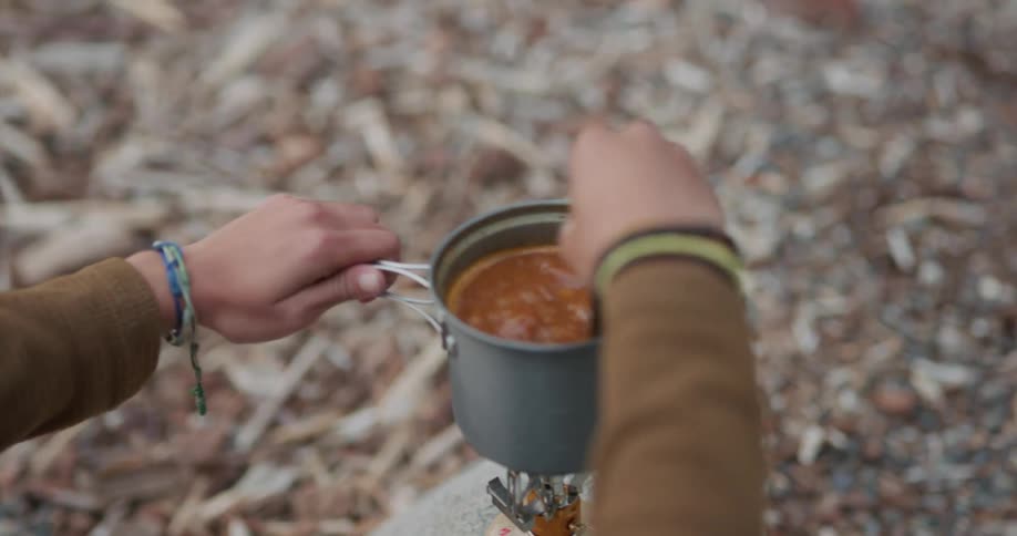 A young woman cooks beans in a small pot at a campsite during the day