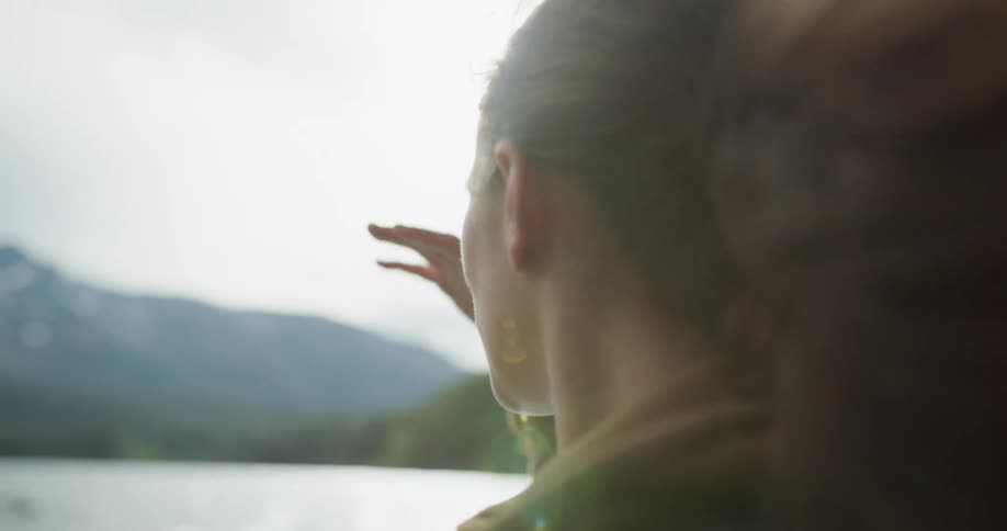 A woman standing on the shore shields her eyes with her hands and looks out across the water