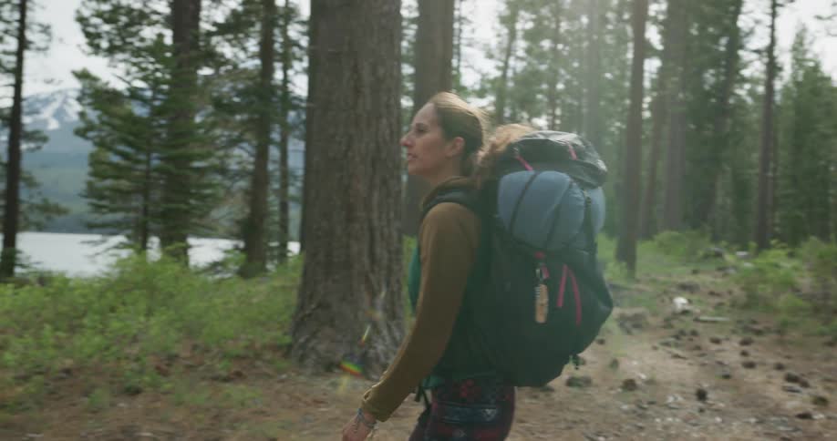 A woman with a backpack walking in a forest next to a lake with the sky and clouds in the background