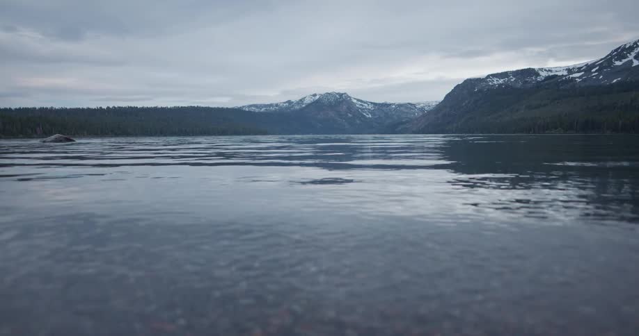 Lake water ripples with snow covered mountains in the distance under a cloudy sky