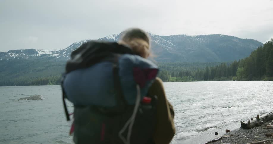 Woman carrying backpack hikes along shore toward forest with mountains on horizon