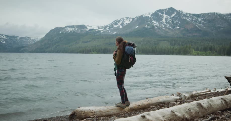 A female hiker standing on a lakeshore with snowy mountains in the distance