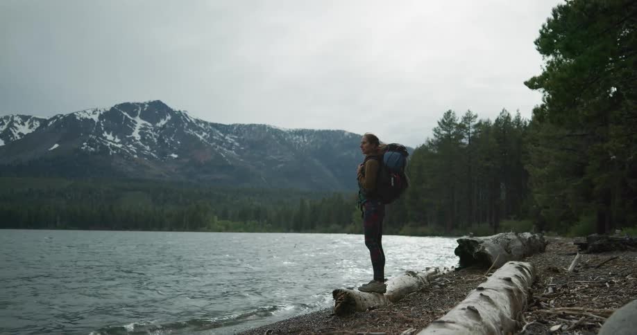 A woman with a backpack standing on a log by a lake with a mountain range the sky and clouds in the background