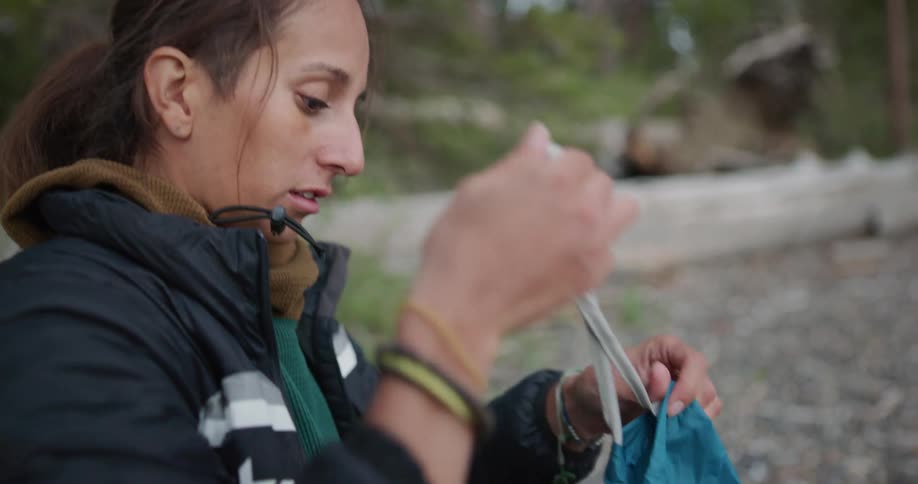 A woman packs a disassembled tent in a carrying bag while kneeling at a campsite during the day