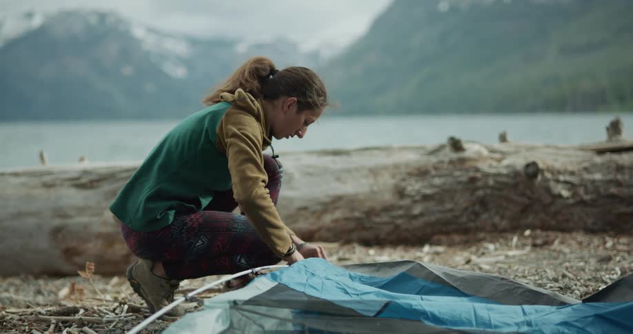 A young woman assembles a tent on the shore of a lake in the wilderness during the day