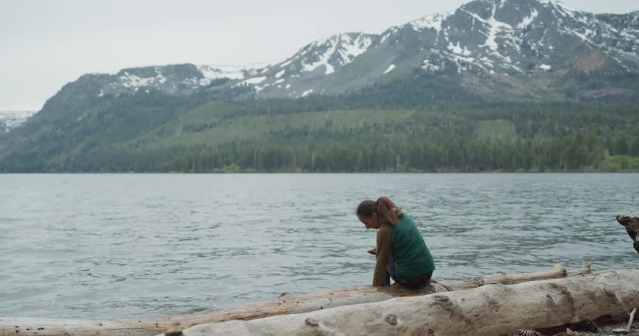 Woman sitting on a log at a lake collecting pebbles from shoreline