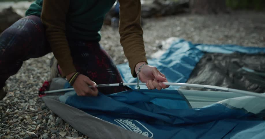 Young woman assembling a tent on the shore of a lake near the pine tree forest in the mountains