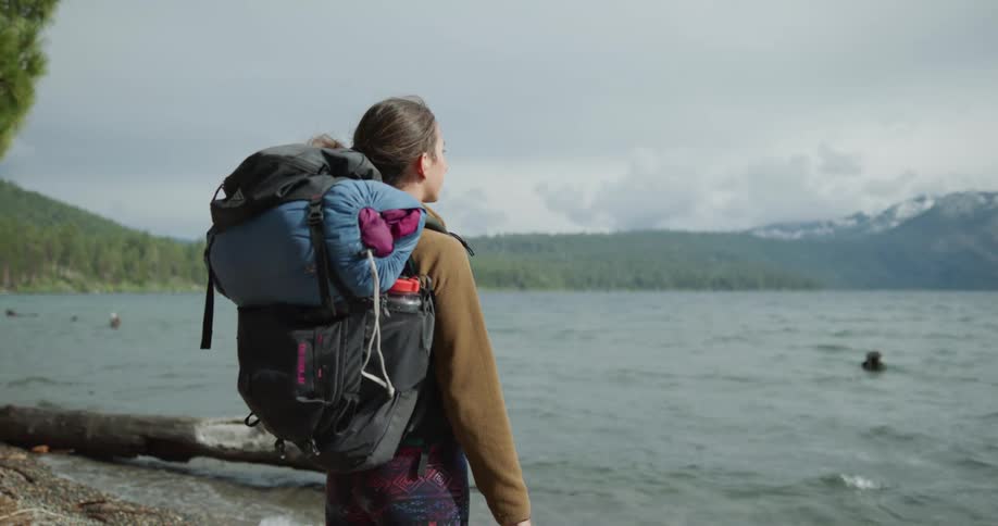 Young woman with a backpack standing on the shore of a lake near the pine tree forest in the mountains