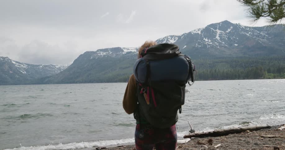 A woman with a backpack walking by a lake with a mountain range the sky and clouds in the background