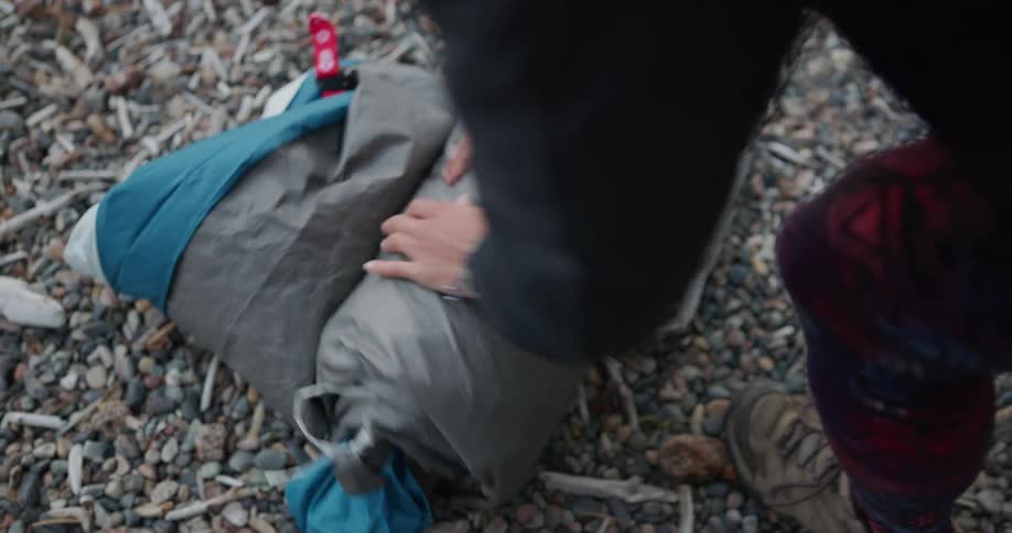 A woman slowly rolls up a sleeping bag at a campsite during the day