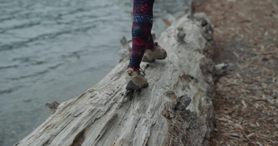 Woman with a ponytail a green brown hoodie and black leggings walks along a tree trunk next to a lake