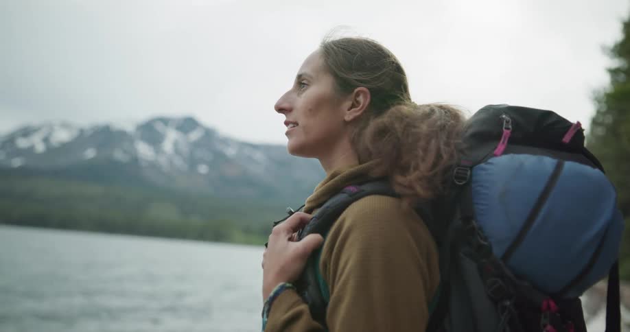 Woman with a ponytail and a backpack stands by a lake on a cloudy day