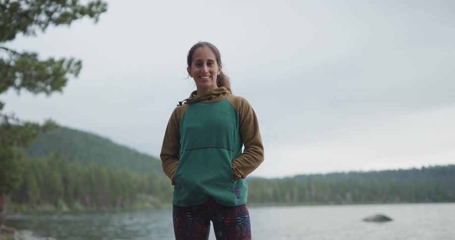 A woman stands posing with hands in her pockets in front of a lake and trees